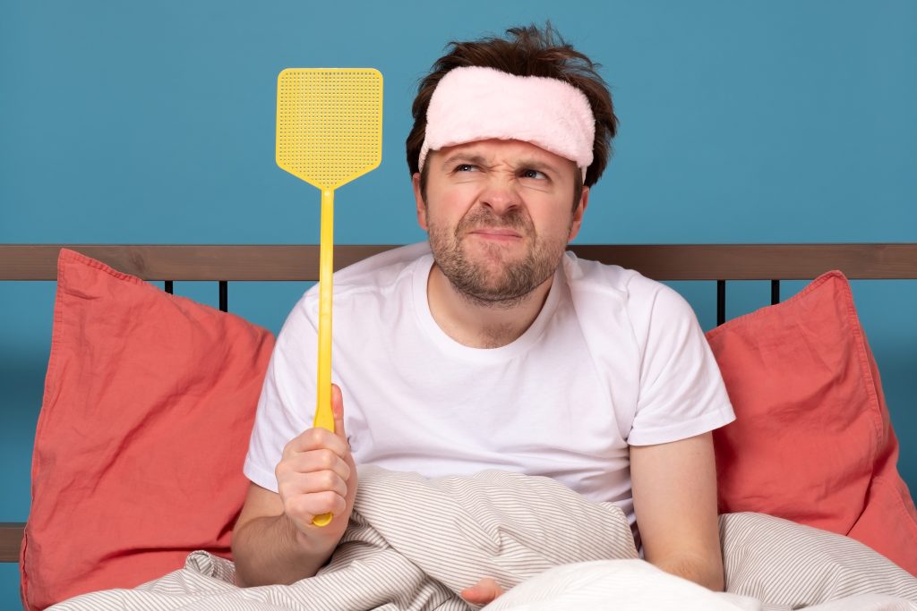 man holding a fly swatter in bed wanting to kill mosquito in bedroom